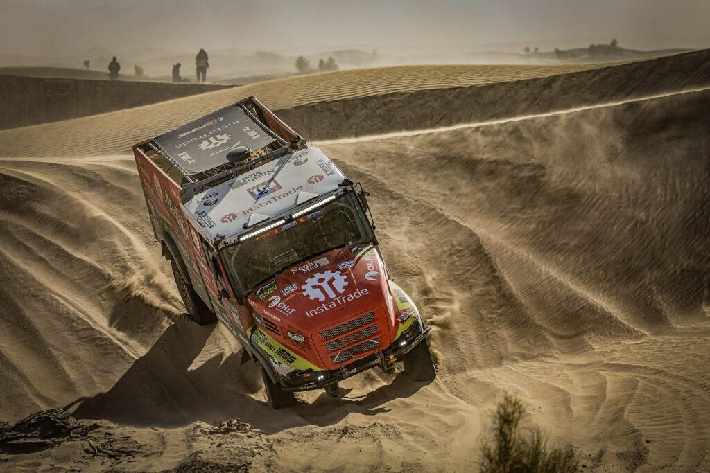 Červený kamion Aleše Lopraise při Rally Dakar 2025, projíždějící duny a vířící prach v terénu Saúdské Arábie; v pozadí siluety diváků.